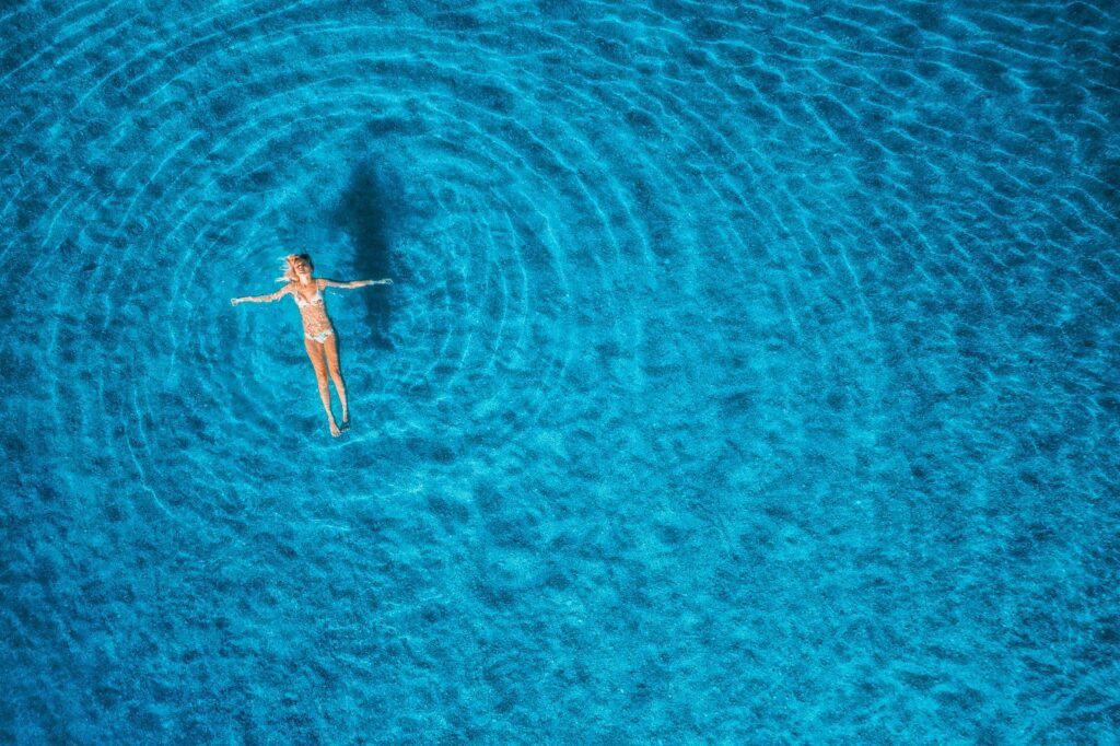 Vue aérienne d'une personne flottant sur le dos dans une eau bleue claire, créant des ondulations dans l'eau, symbolisant la détente et l'évasion en milieu aquatique tropical, parfait pour les articles sur les voyages bien-être.