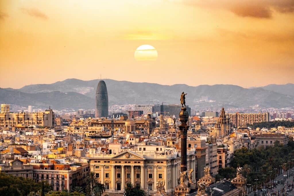 Panorama urbain de Barcelone au coucher de soleil avec la silhouette de la statue de Christophe Colomb en premier plan, la tour Agbar illuminée et les montagnes en arrière-plan, reflétant l'architecture diversifiée et l'histoire riche, parfait pour promouvoir le tourisme culturel en Espagne